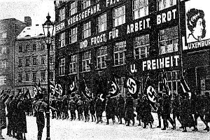 Schwarzes und weißes Bild einer Gruppe von Menschen, die mit Fahnen und Transparenten die Straße entlangmarschieren, mit einem Laternenpfahl links und einer Tafel mit der Aufschrift "Deutsche Nationalsozialistische Arbeiterpartei in Berlin" rechts.