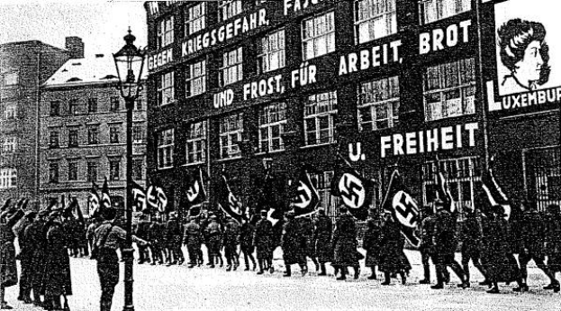 Schwarzes und weißes Bild einer Gruppe von Menschen, die mit Fahnen und Transparenten die Straße entlangmarschieren, mit einem Laternenpfahl links und einer Tafel mit der Aufschrift "Deutsche Nationalsozialistische Arbeiterpartei in Berlin" rechts.
