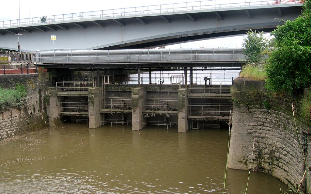 Ein Blick auf die Schleusentore beim Wasserlassen, mit einer Brücke, Bäumen und Pflanzen im Hintergrund.