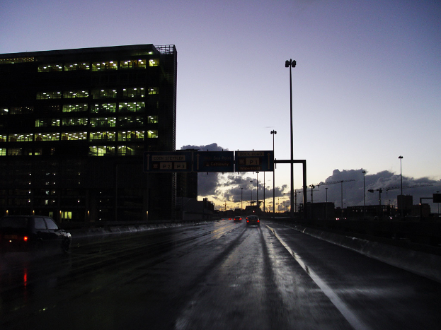 Ein transparentes Gebäude voller Lichter mit mehreren Strommasten und einer Straße mit ein paar Fahrzeugen auf der rechten Seite.