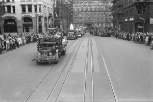Ein Schwarz-Weiß-Foto eines Umzugs auf einer Stadtstraße mit Fahrzeugen, Zuschauern auf beiden Seiten und Gebäuden, Laternenmasten und einem Baum im Hintergrund.