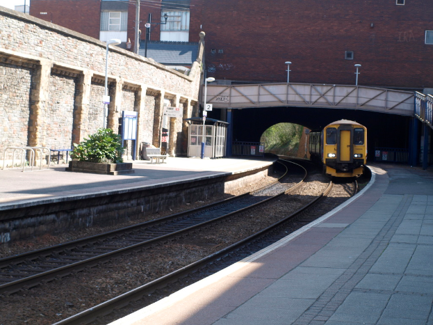 Ein Zug fährt auf den Schienen mit Bahnsteigen auf beiden Seiten, eine Hauswand links, Straßenlaternen, Pflanzen, Bänke, eine Hütte, eine Brücke über die Bahnsteige und Gebäude und Masten im Hintergrund.