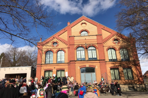 Eine große Gruppe von Menschen steht vor einem Gebäude mit Fenstern und Türen, umgeben von Bäumen unter einem bewölkten Himmel, wahrscheinlich bei einem Stadtfest.