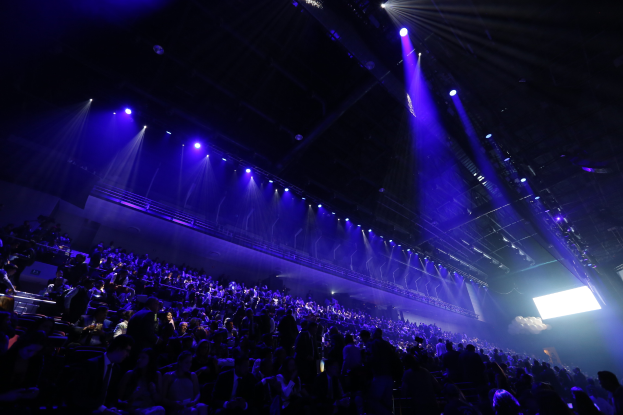Eine große Menschenmenge in einem Konzertsaal vor einer Bühne mit Deckenleuchten und einem Bildschirm auf der rechten Seite.