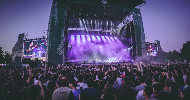 Eine große Menschenmenge auf einem Musikfestival schaut auf einer hell erleuchteten Bühne mit Bildschirmen, die vor einem Gebäude, Bäumen und einem klaren blauen Himmel aufgebaut ist.