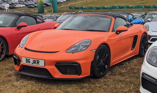 Ein Porsche 718 Boxster GTS auf der Goodwood Festival of Speed mit Autos auf dem Rasen, einem Banner im Hintergrund, Bäumen und einem klaren blauen Himmel.