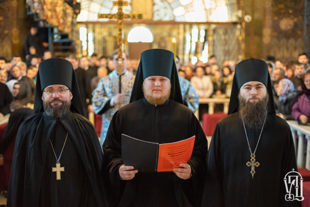 Drei Priester in schwarzen Gewändern und Küppen stehen vor einer Menge in einer Kirche, einer hält ein Buch, mit einem unscharfen Hintergrund und einem Wasserzeichen in der rechten unteren Ecke.