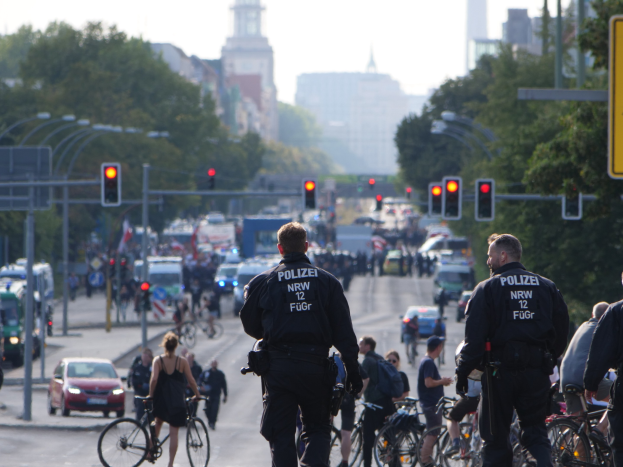 Polizeibeamte fahren mit Fahrrädern eine von Bäumen gesäumte Straße mit Gebäuden und einem klaren Himmel im Hintergrund.