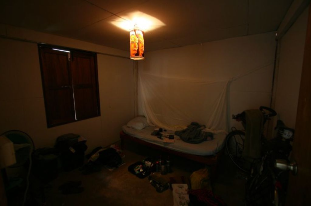 Ein Raum mit einem Bett in der Mitte, ein Fahrrad auf der rechten Seite, ein Fenster auf der linken Seite, ein Tischventilator, Taschen und Flaschen unten, mit Licht von oben.