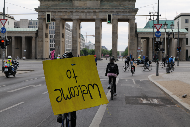 Eine Gruppe von Radfahrern mit Helmen fährt eine Straße vor dem Brandenburger Tor in Berlin, Deutschland, entlang, wobei einer ein gelbes Schild hält, vor einem klaren blauen Himmel mit Gebäuden und Bäumen im Hintergrund.