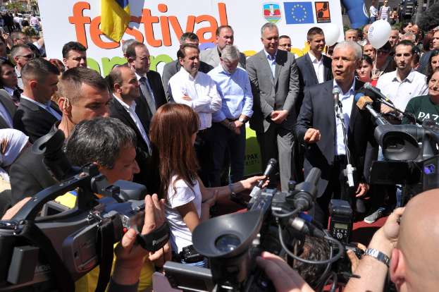 Eine Gruppe von Menschen auf einer Straße mit Kameras und Mikrofonen, mit einem Plakat, Bäumen, Fahrzeugen, Topfpflanzen, einem Ballon, Pfählen und Gebäuden im Hintergrund.