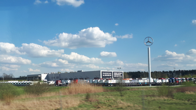 Ein Mercedes-Benz-Werk in Deutschland mit Fahrzeugen auf der Straße, Grünflächen, Gebäuden und einem logo tragenden Pfahl unter einem bewölkten Himmel.