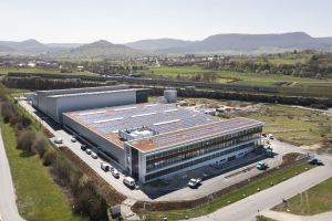 Luftaufnahme eines großen Gebäudes mit Solarmodulen auf dem Dach, umgeben von Fahrzeugen, Bäumen, Gras, Masten und Hügeln unter einem klaren blauen Himmel, identifiziert als Deutschlands erste solarbetriebene Fabrik.