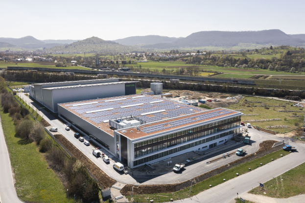 Luftaufnahme eines großen Gebäudes mit Solarmodulen auf dem Dach, umgeben von Fahrzeugen, Bäumen, Gras, Masten und Hügeln unter einem klaren blauen Himmel, identifiziert als Deutschlands erste solarbetriebene Fabrik.