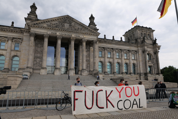 Eine Gruppe von Menschen hält ein Transparent mit der Aufschrift "Fuck You Coal" vor dem Reichstaggebäude in Berlin, Deutschland, mit Bäumen, einem Fahnenmast und einem bewölkten Himmel im Hintergrund.