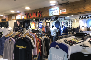 Ein modernes Bekleidungsgeschäft mit zahlreichen T-Shirts auf Ständern und Schaufensterpuppen in aktuellen Outfits, mit beleuchteter Decke und informativen Wandtafeln.