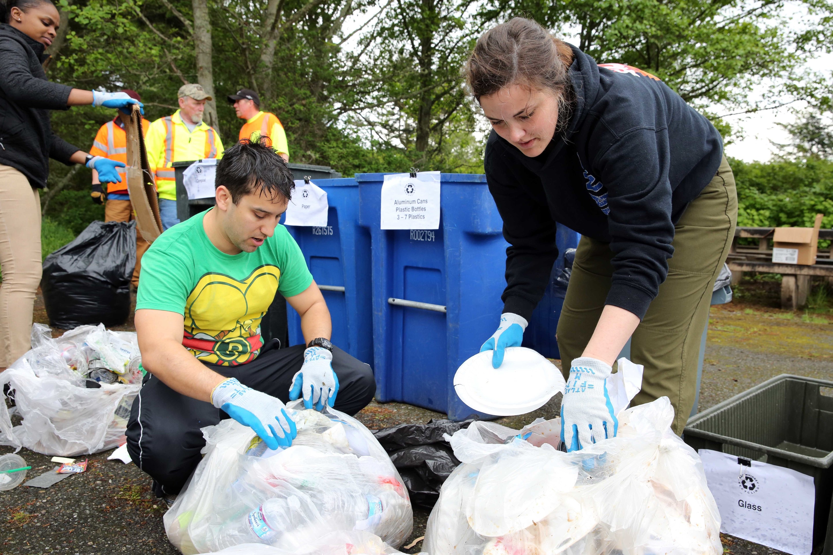 Eine Gruppe von Menschen sammelt Müll in einem Park, mit einem Mann und einer Frau in Handschuhen, die Abfall auf Tellern sammeln, umgeben von Plastik und Flaschen, in der Nähe eines Mülleimers und einer Bank unter Bäumen mit einem klaren Himmel.