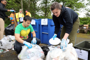 Eine Gruppe von Menschen sammelt Müll in einem Park, mit einem Mann und einer Frau in Handschuhen, die Abfall auf Tellern sammeln, umgeben von Plastik und Flaschen, in der Nähe eines Mülleimers und einer Bank unter Bäumen mit einem klaren Himmel.