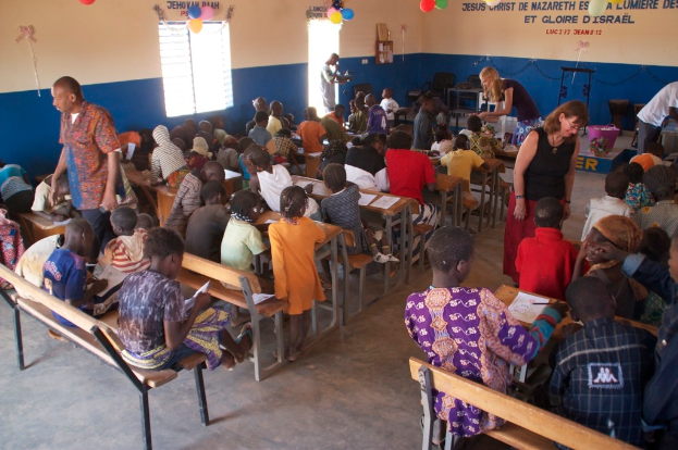 Kinder auf Bänken im Vordergrund, Erwachsene führen sie, Hintergrund mit Stühlen, Tür, Fenster, Tisch, Luftballons und Text oben.