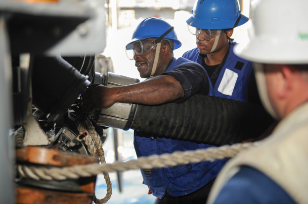 Drei Männer in Helmen und Schutzwesten arbeiten an einer Maschine, mit einem Seil im Vordergrund und einem unscharfen Hintergrund.