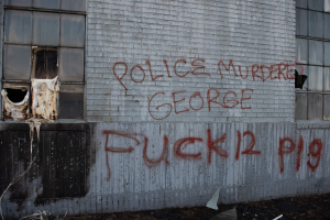 Graffiti auf einer Gebäudewand mit der Aufschrift "Polizeimord George Fuck 12 P19" mit Fenstern und verstreuten Gegenständen davor.