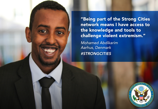 Ein Mann im Anzug und Krawatte, der fröhlich in die Kamera lächelt, mit einem Logo und Text auf der rechten Seite, der besagt: "Teil des starken Städtenetzwerks zu sein bedeutet, Zugang zu Wissen und Werkzeugen zu haben, um gewalttätigen Extremismus in Frage zu stellen", und einem unscharfen Hintergrund.