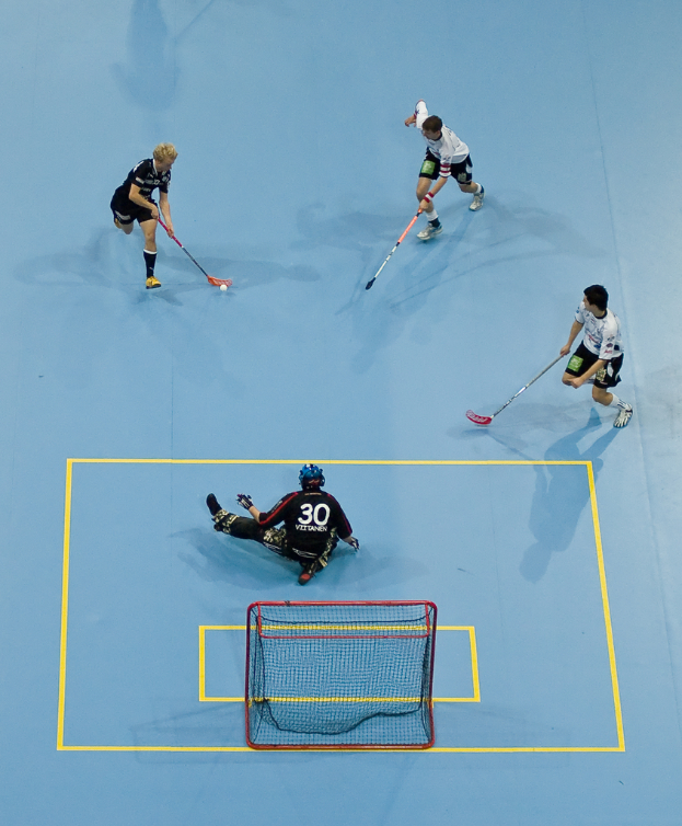 Drei Personen in Sportbekleidung spielen Hockey, wobei eine Person auf der rechten Seite den Ball mit einem Hockeystock schlägt und eine weitere Person hinter einem Tor auf dem Boden sitzt.