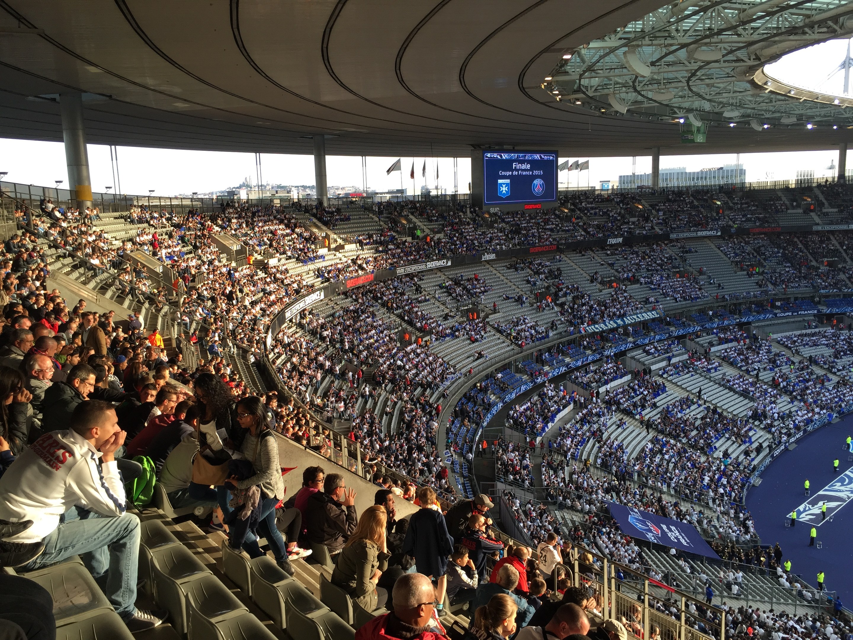 Großes Publikum in der Allianz Arena bei einem Fussballspiel, mit einer Bühne auf der rechten Seite, Fahnen, Masten und einem Bildschirm im Hintergrund unter einem sichtbaren Himmel.