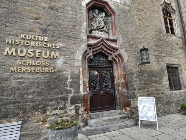 Das Kulturhistorische Museum in Schloss Merseburg, Deutschland, ein Gebäude mit Fenstern, einer Tür, einem Schild, einer Bank, Topfpflanzen und einer Pfeife, unter einem bewölkten Himmel.
