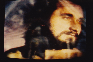 Ein Mann mit langem dunklem Bart und Schnurrbart, der ein weißes Hemd trägt, erscheint auf einem Fernseher mit dunklem Hintergrund und schaut direkt in die Kamera mit einem ernsten Gesichtsausdruck.