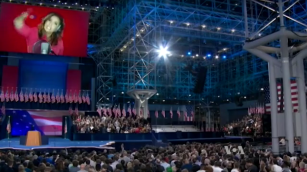 Eine große Menschenmenge sitzt vor einer Bühne mit einem Rednerpult, Mikrofon, Flaggen, einem Bildschirm mit einer Frau und Lichtausrüstung, wahrscheinlich bei einer Präsidenten-Debatte.