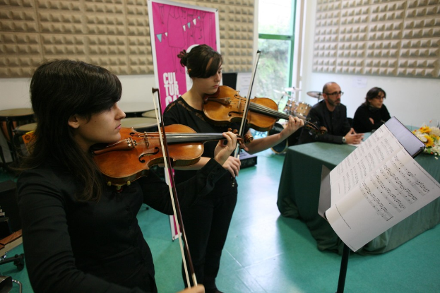 Zwei Frauen, die Geige spielen, eine stehend und eine sitzend, mit einem Notenständer und einem Tisch davor und zwei Personen, die rechts auf Stühlen sitzen, mit einer Tafel im Hintergrund.