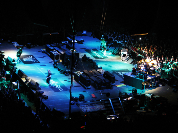 Eine Gruppe von Menschen, die auf einer Bühne mit Lautsprechern, Scheinwerfern und Mikrofonen performen, umgeben von einer großen Menge in einem dunklen Hintergrund.