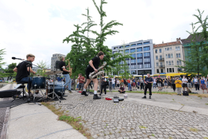 Eine Gruppe von Menschen, die Musik auf einem Bürgersteig mit verstreuter Ausrüstung spielt, vor einem Publikum, mit Bäumen, Gebäuden und einem bewölkten Himmel im Hintergrund.