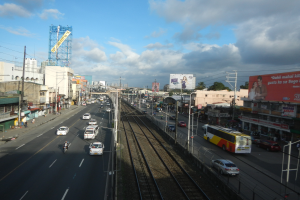 Eine belebte Stadtstraße mit Verkehr, neben einer Bahnschiene, mit Gebäuden, Strommasten, Schildern, Bäumen und einem bewölkten Himmel.