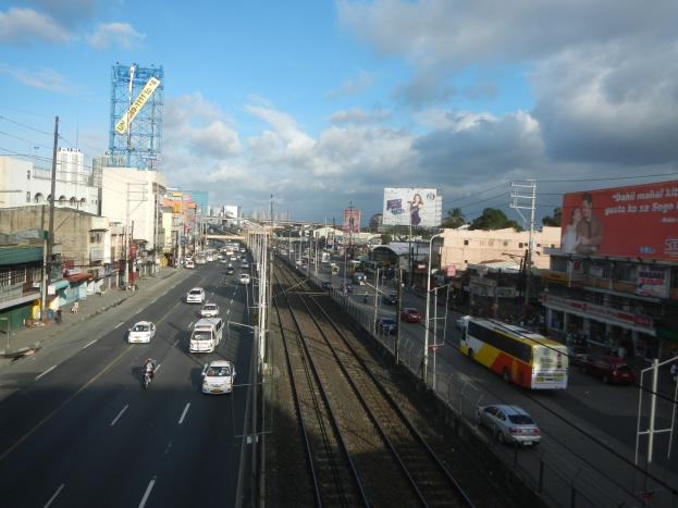Eine belebte Stadtstraße mit Verkehr, neben einer Bahnschiene, mit Gebäuden, Strommasten, Schildern, Bäumen und einem bewölkten Himmel.