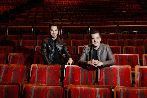 Zwei Männer in schwarzen Jacken, die während einer Vorstellung von *Dancing with the Stars* in einer roten Auditoriumsreihe lächeln.