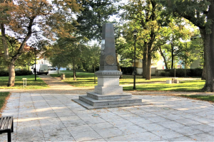 Ein Denkmal steht in einem Park mit einer Bank auf der linken Seite, umgeben von Bäumen, Laternenpfählen und Fahrzeugen auf einer nahen Straße, mit Gebäuden und einem klaren blauen Himmel im Hintergrund.