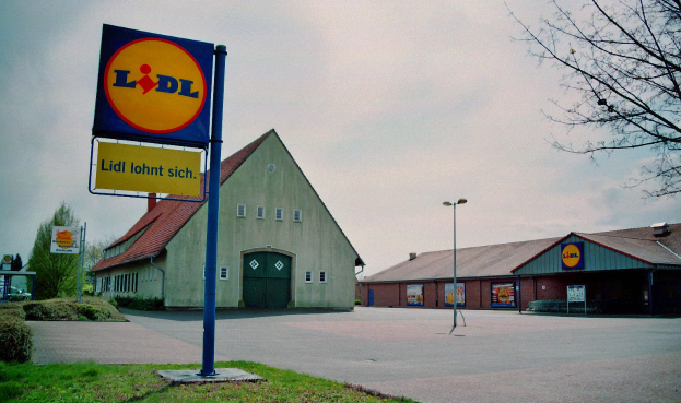 Ein Lidl-Laden mit einem Schild davor, umgeben von Häusern, Straßenlaternen, Autos auf der Straße, Büschen, Bäumen und einem bewölkten Himmel im Hintergrund.