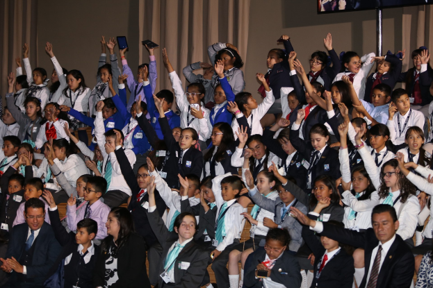 Gruppe von Kindern auf einer Bühne sitzend mit einem Mann und einer Frau davor, Bühne mit Vorhängen und einer Wand dekoriert, Fernseher auf einem Ständer im Hintergrund.