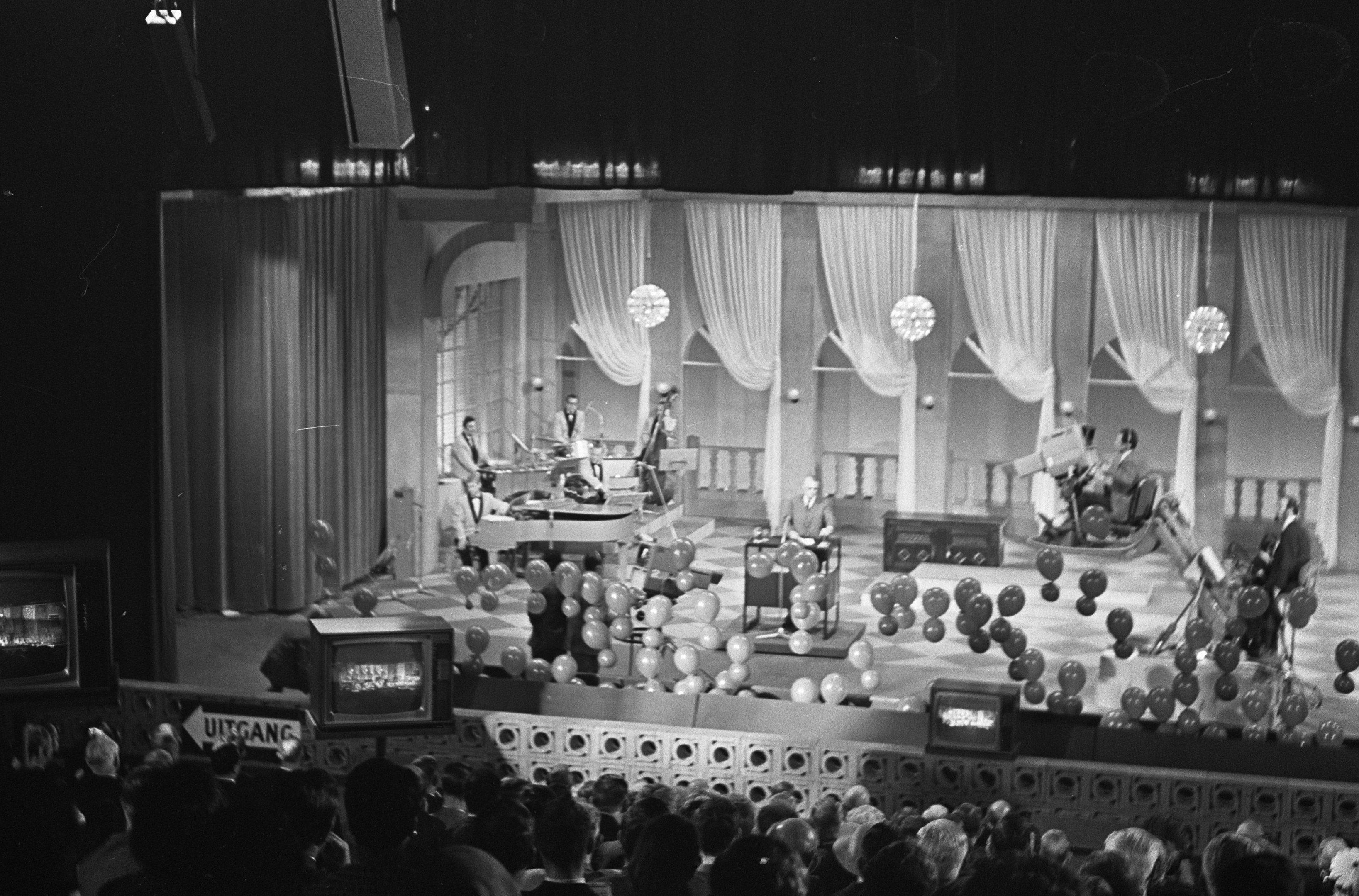 Schwarzes und weißes Bild einer Gruppe von Menschen auf einer Bühne mit Ballons und Fernsehern im Hintergrund.