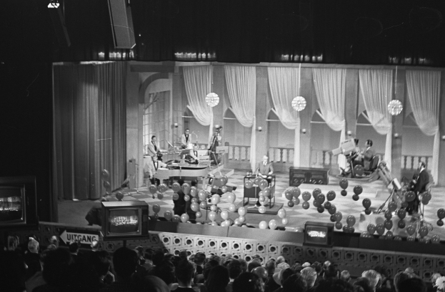 Schwarzes und weißes Bild einer Gruppe von Menschen auf einer Bühne mit Ballons und Fernsehern im Hintergrund.