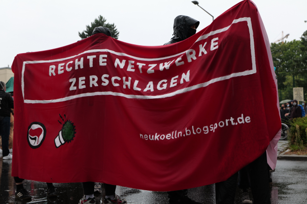 Eine Gruppe von Menschen steht auf einer Straße und hält ein rotes Banner mit der Aufschrift "Rechte Netzwerke Zerschlagen", mit Bäumen, Gebäuden, Laternenpfählen und einem klaren blauen Himmel im Hintergrund, eine Person fährt rechts auf einem Fahrrad und eine weitere Person hält links einen Schirm.