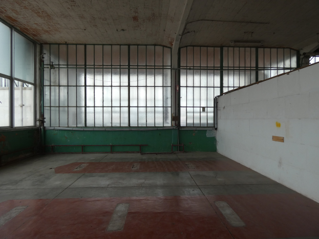 Leerer Raum mit rotem Boden, weißer Wand auf der rechten Seite, Metallstangen und Fenstern im Hintergrund, die an eine Gefängniszelle erinnern.