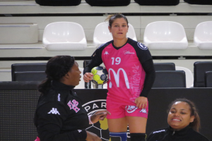 Eine Frau in einem pinken Shirt und Shorts hält einen Fußball, stehend zwischen zwei sitzenden Frauen in einer Stadionkulisse mit Stühlen und einer Wand im Hintergrund.