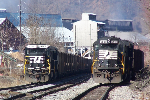 Zwei Züge auf parallelen Gleisen mit Bäumen, Strommasten und Gebäuden im Hintergrund.