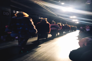 Eine Gruppe von Menschen läuft über den Laufsteg bei einer Fashion Show, beleuchtet von hellen Scheinwerfern, mit Zuschauern auf beiden Seiten.