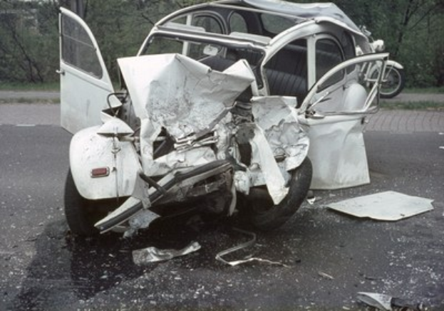 Ein schwer beschädigtes Auto mit Frontalcrash am Straßenrand, umgeben von Bäumen und einem Gebäude, mit einem anderen Fahrzeug im Hintergrund.