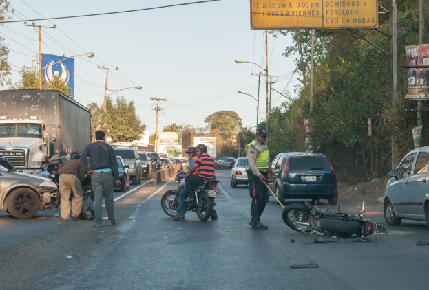 Eine Gruppe von Menschen steht um ein verunglücktes Motorrad auf der Straße mit mehreren Fahrzeugen, darunter ein Lastwagen, und einem Hintergrund aus Bäumen, Polen, Lampen und Schildern unter dem Himmel.