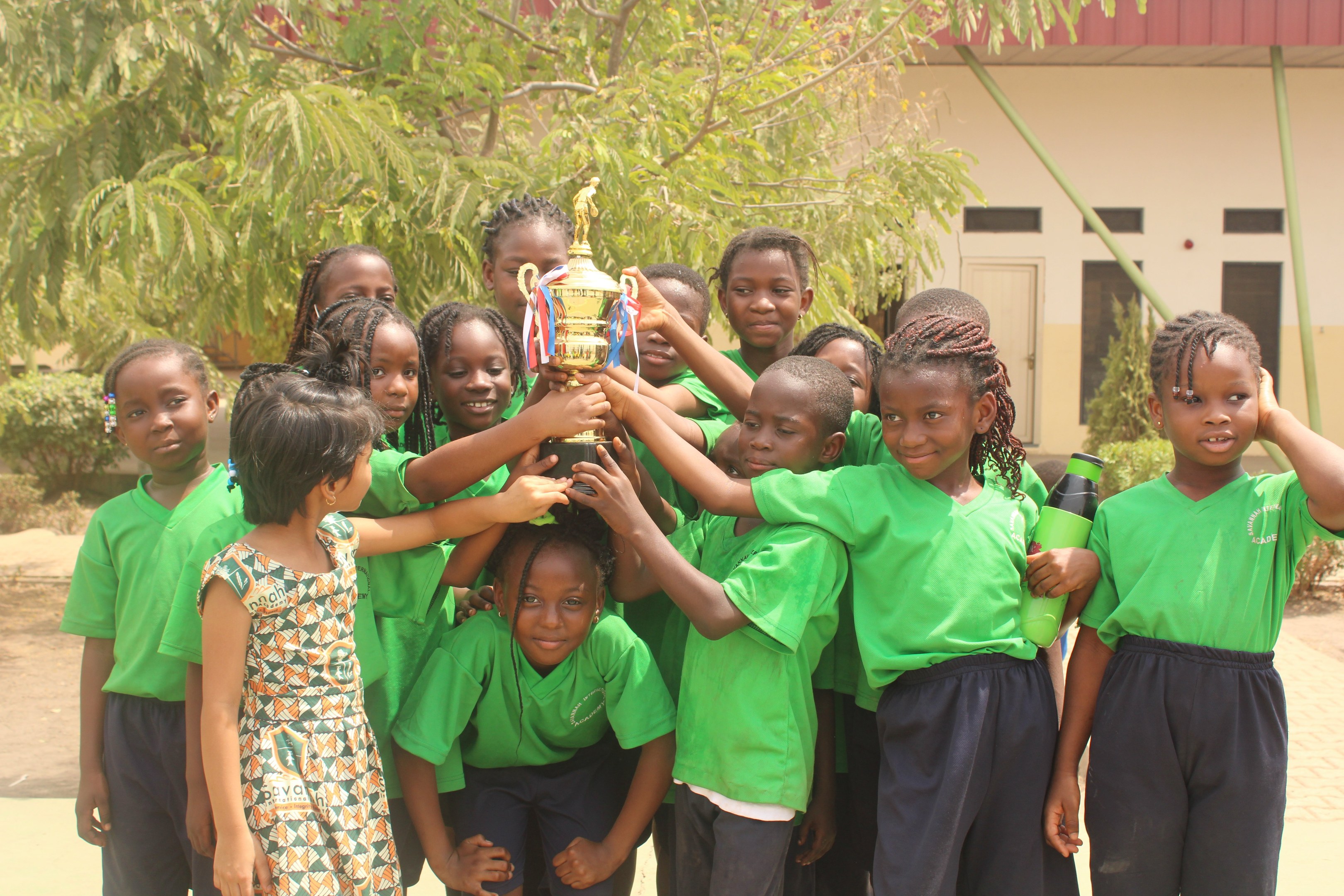 Gruppe von Kindern, die eine Trophäe in einer Schulumgebung mit Bäumen und einem Gebäude im Hintergrund halten.